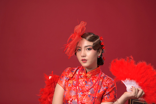 Beautiful asian woman wearing red traditional chinese dress sitting holding feather fan looking to camera on red background, Thailand