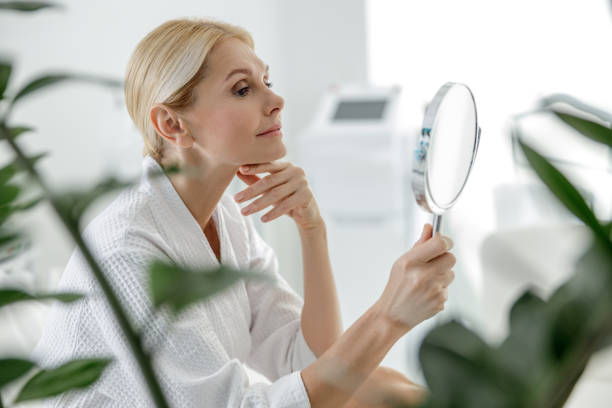 Smiling pretty woman using mirror in spa center Side view of attractive female enjoying time in beauty salon stock photo bathrobe photos stock pictures, royalty-free photos & images