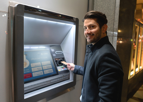 Portrait of a happy man withdrawing money at a cash point and looking at the camera holding his debit card. **DESIGN ON SCREEN AND CREDIT CARD WERE MADE FROM SCRATCH BY US)