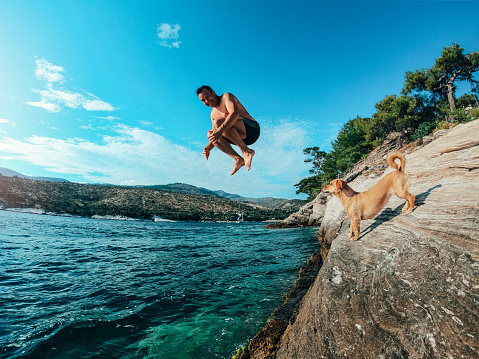 Men jumping into the sea from the rocky shore while his small yellow dog standing on the beach and looking at him