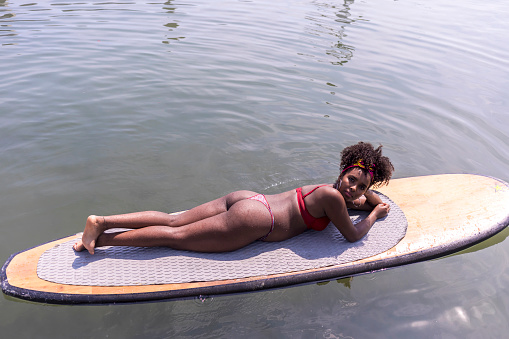 Young afro slim woman lying on a paddle Board in sea.