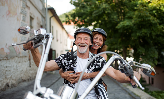 A front view of cheerful senior couple travellers with motorbike in town.