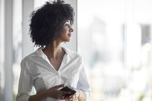 Profile view of contented Brazilian businesswoman in mid 30s with curly black hair and casual open collar white blouse looking out office window.