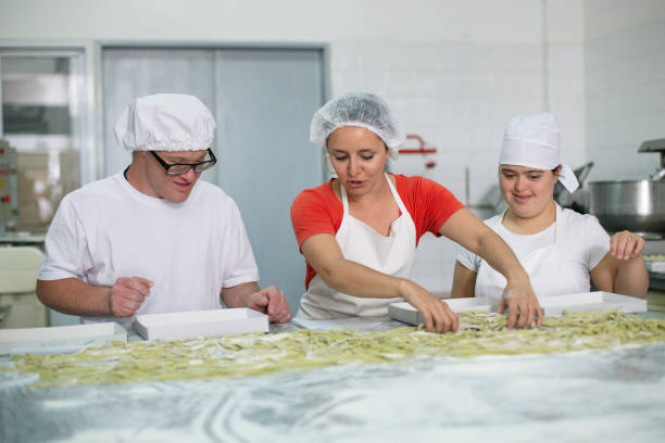 Female Supervisor Training Pasta Makers in Factory Smiling male and female coworkers with Down Syndrome listening to instructions from supervisor about how to organize freshly made fettuccine in boxes. down syndrome photos stock pictures, royalty-free photos & images