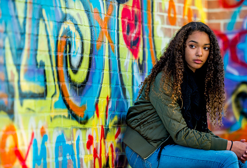 A female African American hipster student, with long dark curly hair, sits against a brick wall covered in graffiti.  She is dressed casually and wearing a fall coat as she holds her cell phone in her hand.