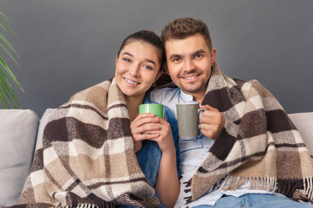 istock Relationnship Concept. Young couple sitting on sofa hugging covered with blanket studio isolated on grey drinking hot tea smiling happy to camera 1193990867