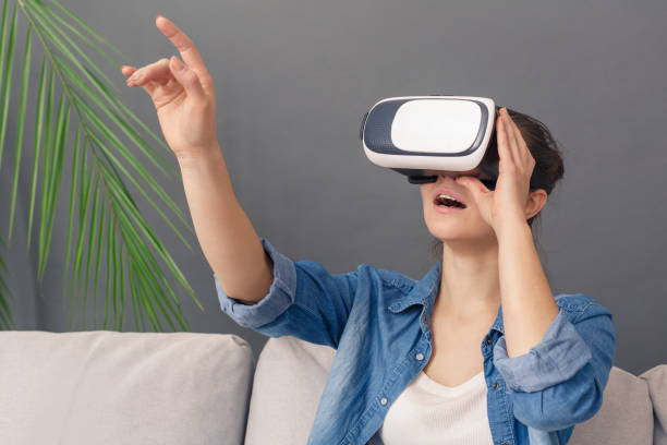 istock Freestyle. Young woman in virtual reality headset sitting on sofa studio isolated on grey watching video smiling impressed 1193990872