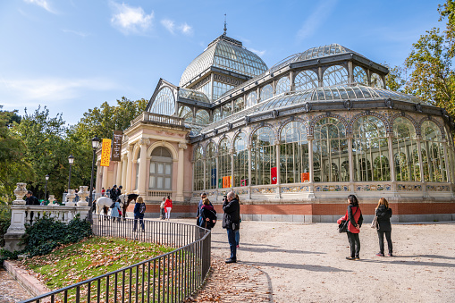 Madrid / Spain - October 16, 2019: People explore El Retiro Park, stopping to enjoy the view at the famous landmark, the Crystal Palace or Palicio de Cristal, an art gallery overlooking a pond.