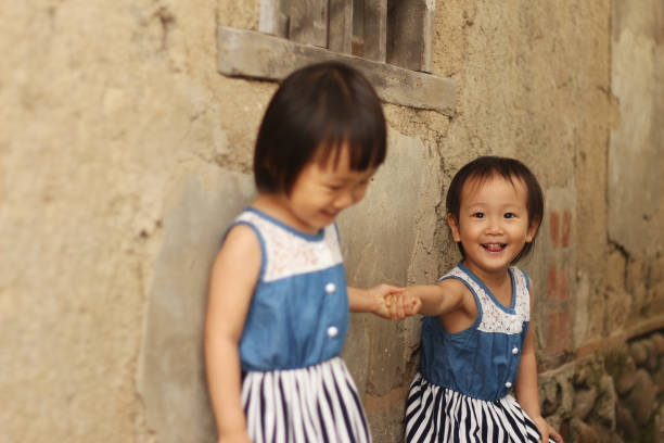 two Asian twins girls hand in hand walking and playing in front of a old house with mottled olden wall