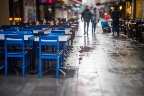 Pedestrians walk between restaurants or cafes on a narrow, rainy street in Europe