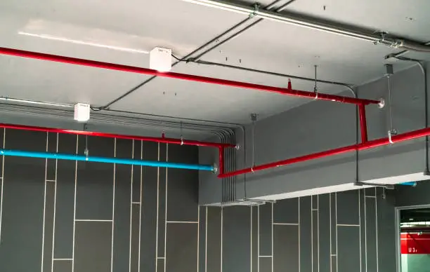 Photo of Automatic fire sprinkler safety system and red water supply pipe. Fire Suppression. Fire protection and detector. Fire sprinkler system with red pipe hanging from ceiling inside building. Wiring tube.