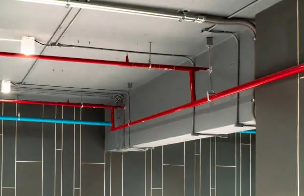 Photo of Automatic fire sprinkler safety system and red water supply pipe. Fire Suppression. Fire protection and detector. Fire sprinkler system with red pipe hanging from ceiling inside building. Wiring tube.