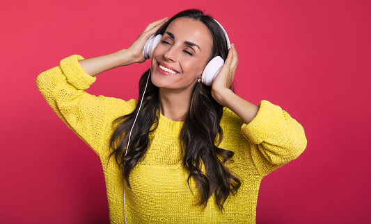 Close-up photo of a wonderful young girl with long dark hair in white headphones, who is listening to music with her eyes closed, smiling and holding them with her hands.