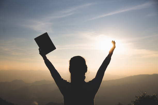 mujeres de pie sosteniendo la santa biblia para adorar a dios en el fondo de la puesta del sol, orar al dios, concepto de silueta cristiana. - god freedom arms raised high angle view fotografías e imágenes de stock