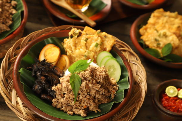 Nasi Megono, traditional Javanese rice dish from Pekalongan, Central Java. It is steamed rice with Megono (stir-fried minced young jackfruit) as the main side dish; then added with Cumi Hitam (braised squid in its ink), Pindang Telur (braised egg in soy sauce) and Tempe Mendoan (fried tempeh in batter). The dish is plated on an earthenware plate lined with banana leaf. Served with Sambal (chili sauce) as the condiment. The Nasi Megono and Sambal are arranged on a table with extra portion of Tempe Mendoan.