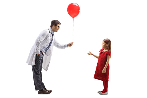 Full length profile shot of a male doctor giving a red balloon to a girl isolated on white background