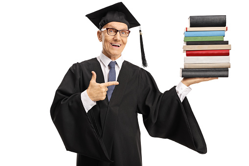 Senior man wearing a graduation gown and holding a pile of books isolated on white background