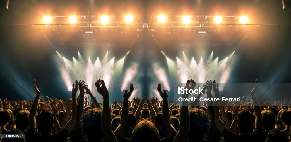 Concert stage, people are visible waving and clapping, silhouettes are visible A shot taken in front of a concert stage lit in the night, people are visible waving and clapping, but no one is recognizable. Music Festival Stock Photo