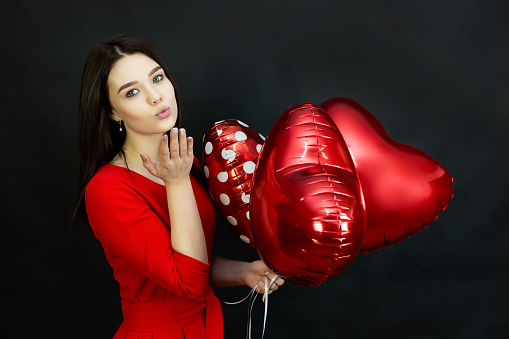 Teenager girl in a red dress with inflatable balloons blows a kiss on a black background.