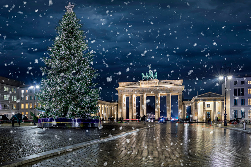 Panoramic view to the famous Brandenburg Gate in Berlin, Germany, with a illuminated Christmas tree in front and falling snow during winter night time
