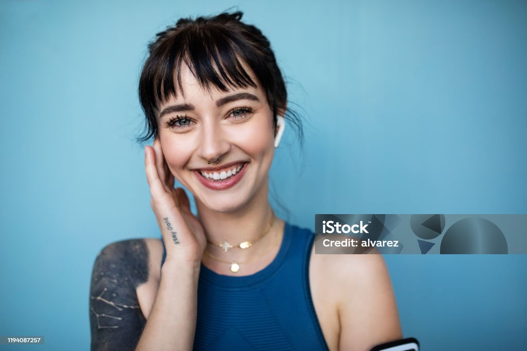 Fitness woman listening to music on wireless headphones Close-up portrait of beautiful woman listening to music on her wireless headphones against blue background. Cheerful young female in sportswear looking at camera. Women Stock Photo