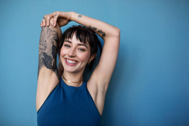 Beautiful young woman posing in sportswear Studio shot of a beautiful young woman posing in sportswear against a blue background. Smiling woman in sportswear with tattoo looking at camera. tattoo photos stock pictures, royalty-free photos & images