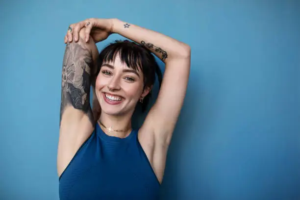 Studio shot of a beautiful young woman posing in sportswear against a blue background. Smiling woman in sportswear with tattoo looking at camera.