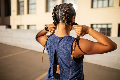 Rear view of young woman standing outdoors holding her braided hair. Fit woman in sports wear working out in city.