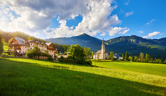 Austria mountain landscape. Traditional church chapel village. Scenic evening sunset panoramic view in austrian Alps mountains. Summer green lawns fields. Knolls covered forest trees. Blue sky clouds.