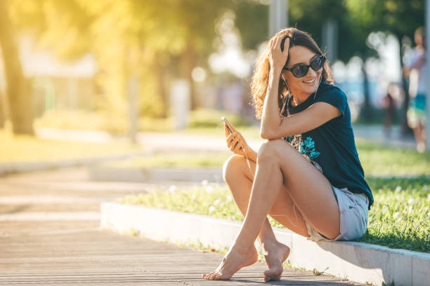 giovane bella donna felice seduta sul prato con smartphone e cuffie che ascoltano musica o audiolibri - parlare e ridere sul telefono cellulare - calda immagine del tramonto estivo - main street audio foto e immagini stock