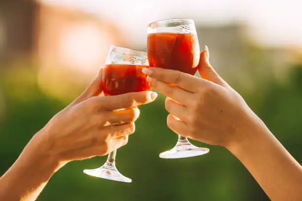 Photo of Cheers. Female hands toasting on sunset with red wine fresh sangria glasses with green sunny garden background - female friendship, holidays celebration and party concept