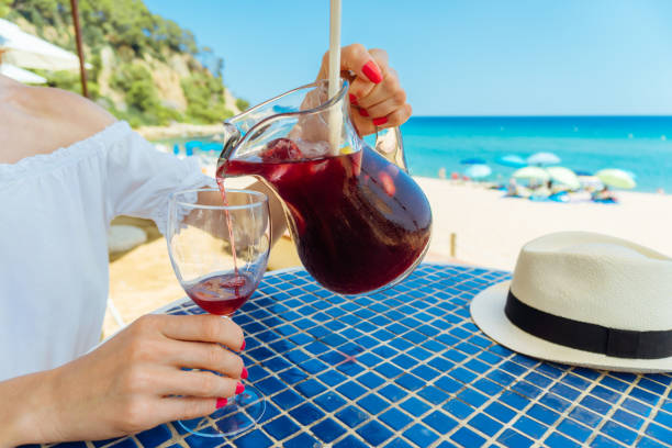 imagem de bela turista hipster derramando sangria de vinho tinto fresco no copo sentado em um café de praia (chiringuito) - red jug - fotografias e filmes do acervo