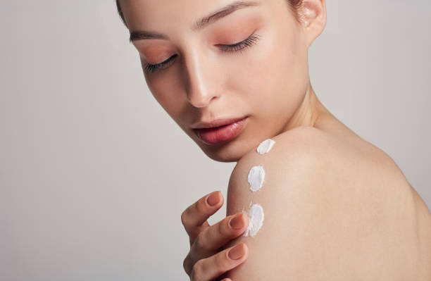A young girl with clean fresh skin is applying cream on her shoulder with smears. Portrait on a light background. Beauty and care concept. stock photo