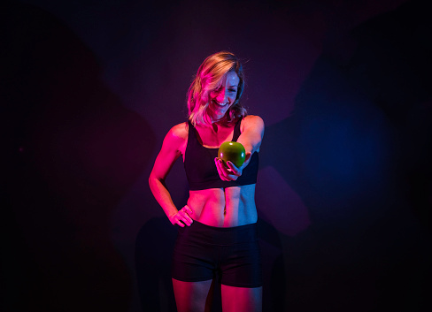 One person / waist up / front view of 20-29 years old adult beautiful caucasian young women / female standing in front of black background / multi-colored background / colored background wearing sports clothing / sports bra / shorts / running shorts who is smiling / happy / cheerful / black color / gel effect lighting / apple - fruit / orange - fruit / banana / grape