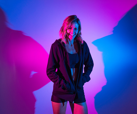 Waist up / one person of 20-29 years old adult beautiful caucasian female / young women standing in front of multi-colored background / colored background wearing sports clothing / sports bra / shorts / running shorts / jacket who is smiling / happy / cheerful with shadow / gel effect lighting
