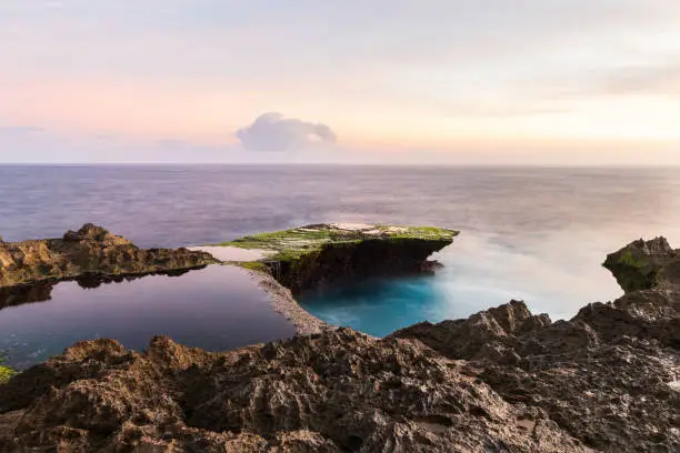 Devil's Tear at sunset, island of Nusa Lembongan, Bali, Indonesia. Rocky shore in foreground. Yellow sky with clouds beyond.