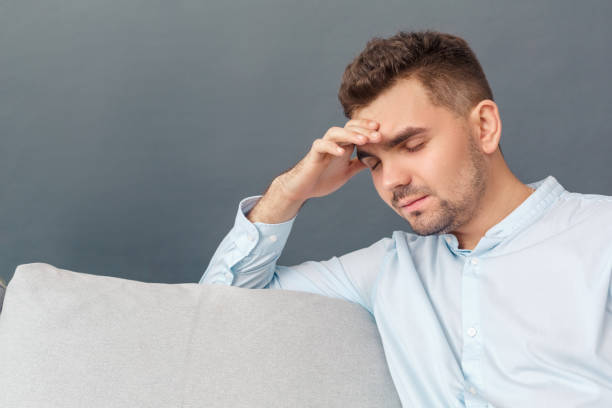 istock Freestyle. Young man studio sitting on sofa isolated on grey touching head having headache tired close-up 1194177718
