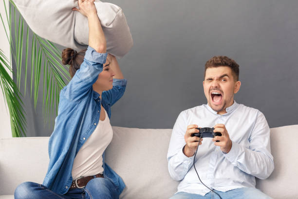 istock Relationnship Concept. Young couple woman hitting with pillow man playing game console screaming sitting on sofa studio isolated on grey 1194177721
