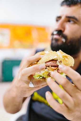 Close up portrait of bearded man eating burger. Portrait of expressive fat man eating hamburger with meat, tomato, bacon, onion and cheese. Cheeseburger and fast food concept.