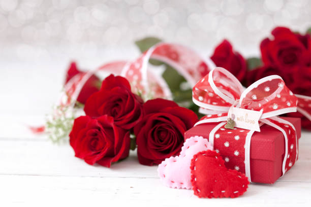 Close-up view of a  wooden calendar  highlighting February 14, surrounded by a romantic bouquet of red roses. The warm and vibrant setup is ideal for Valentine's Day visuals and romantic-themed designs,On paper red and pink hearts with a red gift box,