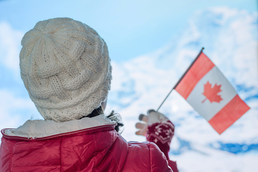 Back view of young woman in a winter knitted white cap, red jacket against the backdrop of the snowy mountains. Girl with Canadian flag on the top of mount