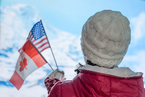 Back view of young woman in a winter knitted white cap, red jacket against the backdrop of the snowy mountains. Girl with American Canadian flags on the top of mount