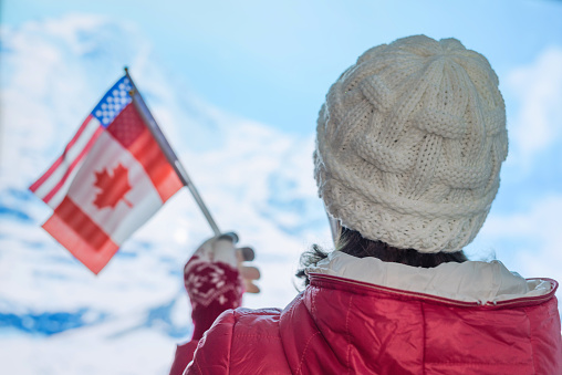 Back view of young woman in a winter knitted white cap, red jacket against the backdrop of the snowy mountains. Girl with American Canadian flags on the top of mount
