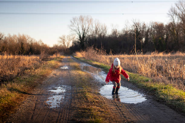3 years old child jumping in a puddle with red jacket and wellies on a sunny winter day. stock photo