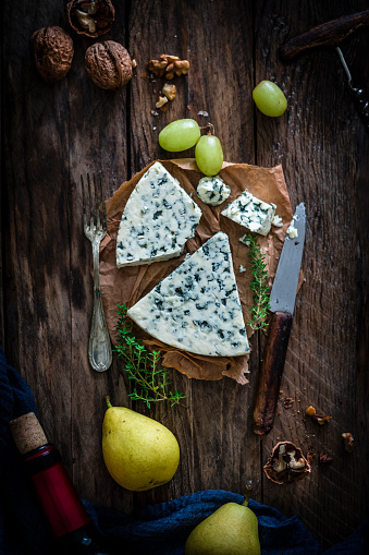 Blue cheese slices shot from above on rustic wooden table. The cheese slices are sitting on a brown paper piece. A kitchen knife, walnuts, grapes, pears and a wine bottle complete the composition. Predominant colors are white and brown. High resolution 42Mp studio digital capture taken with Sony A7rii and Sony FE 90mm f2.8 macro G OSS lens
