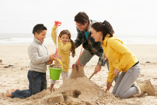 Blond toddler boy play building sand castle on the beach and happy mother laugh on background