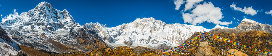 Deep blue high altitude skies over the snow capped peaks and dramatic pinnacles of Huinchuli (6441m), Annapurna South (7219m) and Annapurna I (8091m) towering above the Buddhist prayer flags flying at Annapurna Base Camp deep in the remote mountain wilderness of the Annapurna Sanctuary, Nepal.