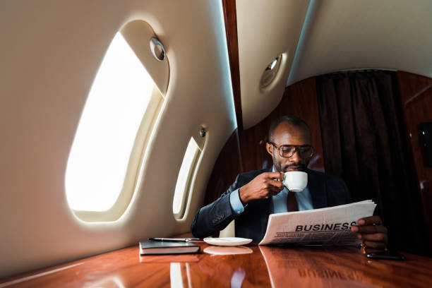 hombre de negocios afroamericano leyendo periódico de negocios mientras bebe café en avión privado - wealthy fotografías e imágenes de stock