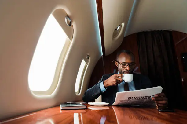 African american businessman reading business newspaper while drinking coffee in private plane