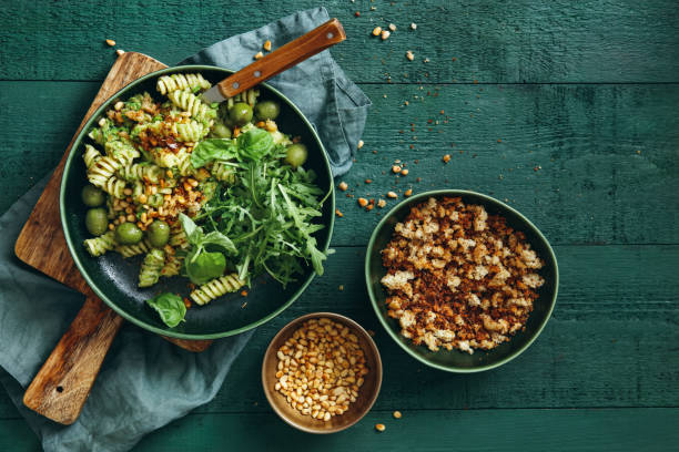 Summer vegetarian pasta salad with broccoli pesto Summer vegetarian pasta salad with broccoli pesto, peas, arugula, olives, pine nuts and bread crumbs on dark green background. Top view. vegan food photos stock pictures, royalty-free photos & images
